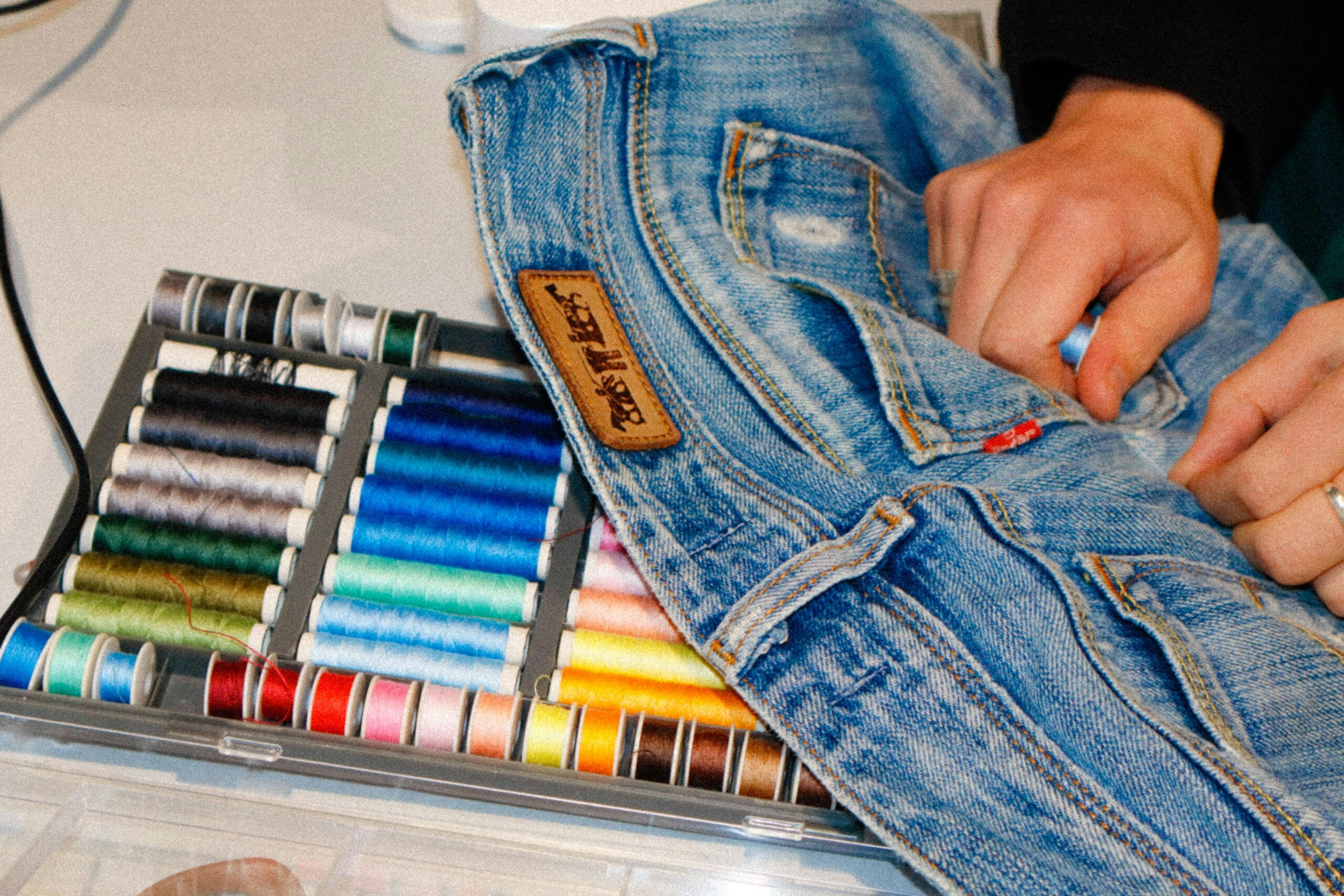 Close-up of hands sewing blue jeans with a variety of colorful threads, showcasing fashion craftsmanship.